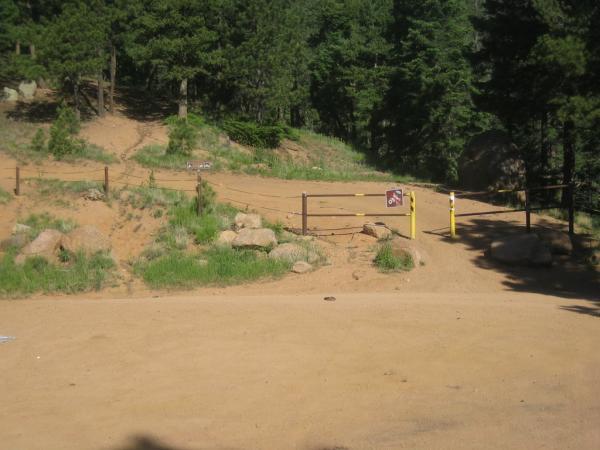 A dirt path leading into a wooded area, with a wooden fence and a gate marked by a sign. The scene is surrounded by green grass and rocks, under a clear blue sky. The dense trees in the background create a natural and serene atmosphere. Bear Creek Canyon Loop mountain bike trail.