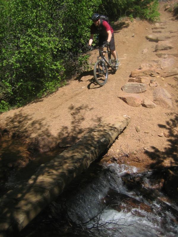 A mountain biker navigating a rocky trail beside a small stream, with vibrant green foliage in the background. The rider is wearing a helmet and riding gear, and a log bridge crosses over the water. Bear Creek Canyon Loop mountain bike trail.