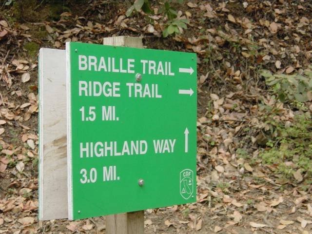 Green trail sign indicating directions for Braille Trail, Ridge Trail (1.5 miles), and Highland Way (3.0 miles), mounted on a wooden post with a natural background of leaves and greenery. Forest Of Nisene Marks and Soquel Demonstration Forest mountain bike trail.