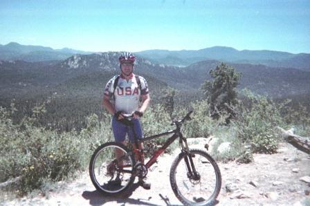 A person wearing a cycling jersey with "USA" printed on it stands next to a mountain bike on a rocky trail, surrounded by a scenic view of mountains and trees. The sky is clear and blue, indicating a sunny day. Elk Meadow mountain bike trail.