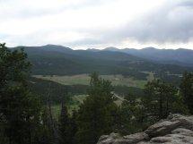 A scenic view of a mountainous landscape, featuring rolling hills, dense forests, and a cloudy sky, taken from a rocky vantage point. 3 Sisters / Alderfer mountain bike trail.