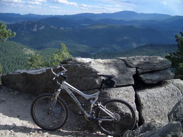 A mountain bike resting on a rock ledge with a panoramic view of lush green hills and distant mountains under a partly cloudy sky. Elk Meadow mountain bike trail.