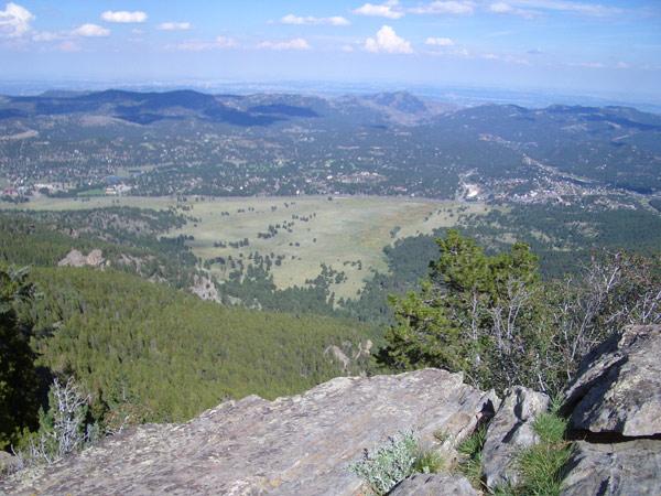 A panoramic view of a lush green valley surrounded by rolling hills and distant mountains, taken from a rocky vantage point. The foreground features rocky terrain with sparse vegetation, while the expansive landscape below showcases a mix of meadows and wooded areas under a clear blue sky with scattered clouds. Elk Meadow mountain bike trail.