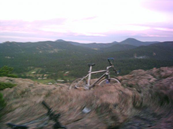 A mountain bike positioned on rocky terrain, overlooking a valley and distant mountains during twilight. The sky displays soft hues of purple and pink, creating a tranquil and scenic outdoor setting. 3 Sisters / Alderfer mountain bike trail.