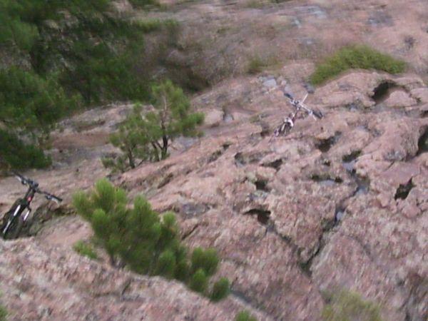 Two mountain bikes are positioned on rocky terrain, surrounded by sparse vegetation and trees. The rocky surface shows textures and crevices typical of a natural landscape. 3 Sisters / Alderfer mountain bike trail.