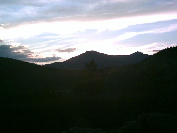 Silhouette of mountains against a sunset sky, with clouds and a gradient of colors transitioning from orange to blue. The foreground features dark outlines of trees and rocky terrain. 3 Sisters / Alderfer mountain bike trail.