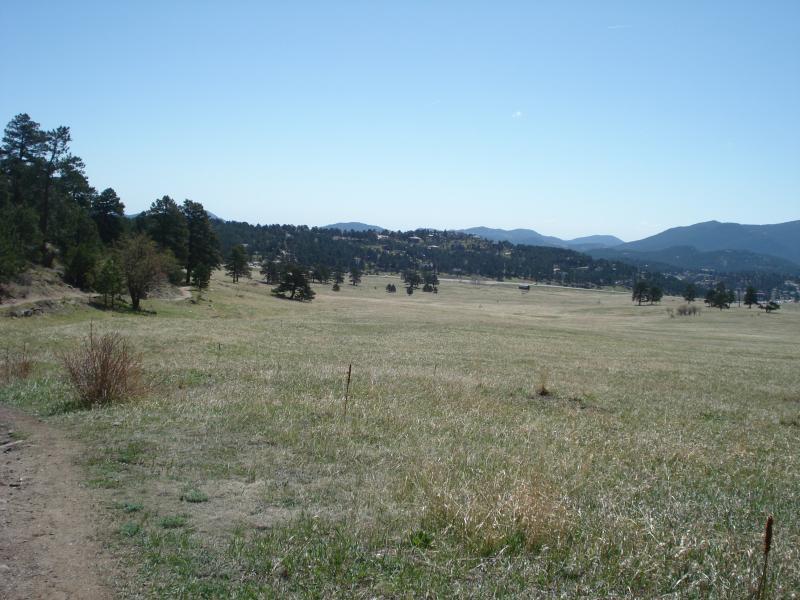 A wide-open grassy field bordered by trees, with distant mountains under a clear blue sky. A dirt path leads into the scene, extending through the foreground. Elk Meadow mountain bike trail.