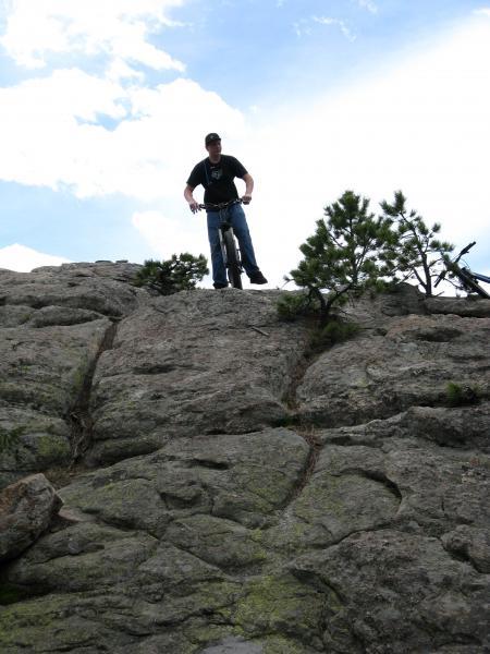 A mountain biker standing on a rocky terrain, poised on a steep incline. The sky is partly cloudy, with patches of blue visible. Sparse vegetation, including small trees, is present among the rocks. The biker is wearing a black T-shirt and jeans, preparing to navigate down the rocky surface. 3 Sisters / Alderfer mountain bike trail.