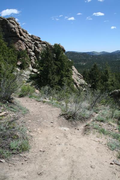 A dirt trail winding through a rocky landscape, flanked by green shrubs and trees, leading to a scenic view of distant mountains and a blue sky with scattered clouds. 3 Sisters / Alderfer mountain bike trail.