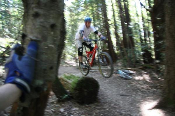 A mountain biker in a white jersey and blue helmet is mid-air, jumping over a log on a wooded trail, with trees and dappled sunlight in the background. A hand wearing a glove reaches out from the left side of the image, appearing to touch a nearby tree trunk. Forest Of Nisene Marks and Soquel Demonstration Forest mountain bike trail.