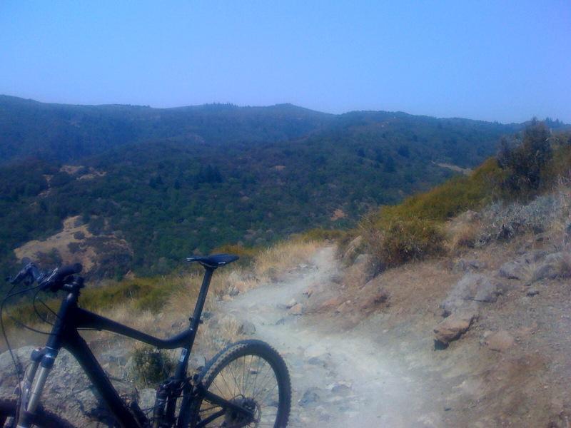 A mountain bike parked on a dirt trail overlooking a scenic mountainous landscape with greenery and hills under a clear blue sky. Camp Tamarancho mountain bike trail.