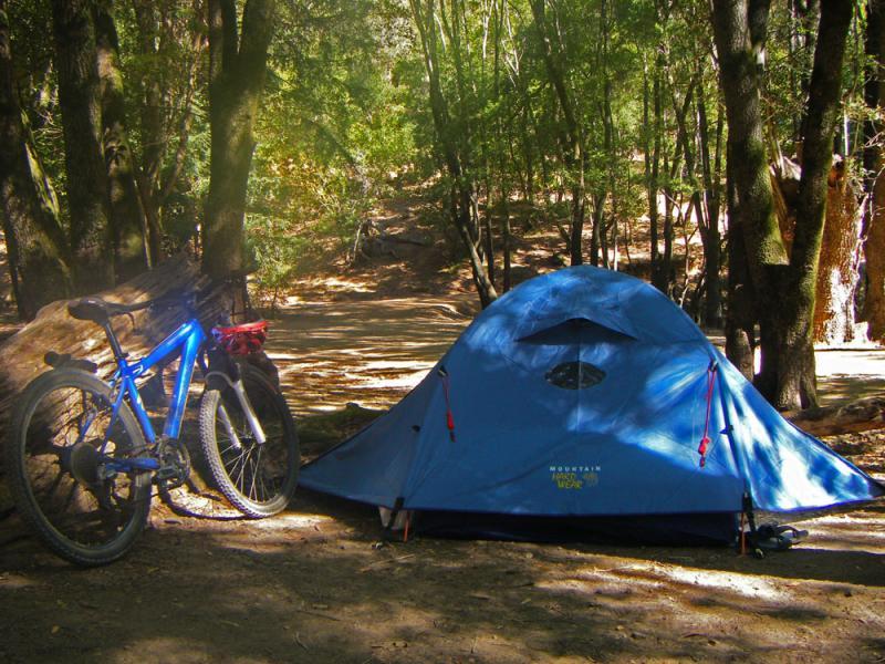 A blue tent set up in a forested camping area, next to a mountain bike. The scene features tall trees and a dirt path in the background, suggesting a peaceful outdoor environment. China Camp mountain bike trail.