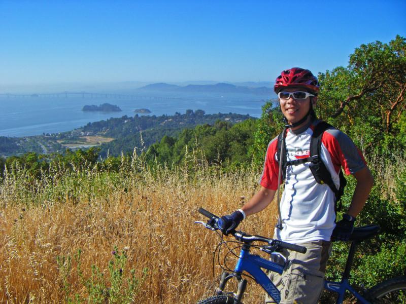 A person wearing a red helmet and sunglasses stands next to a blue mountain bike, smiling at the camera. They are on a grassy hillside with a scenic view of a bay and distant mountains in the background, under a clear blue sky. China Camp mountain bike trail.