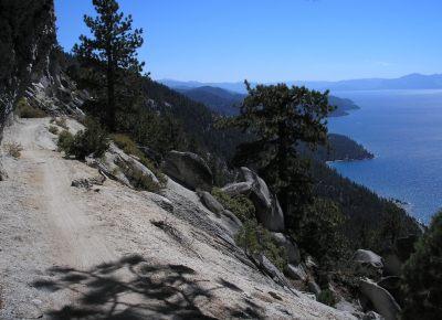A scenic view of a dirt path winding along a cliffside, surrounded by trees overlooking a blue lake and distant mountains under a clear sky. Flume Trail mountain bike trail.
