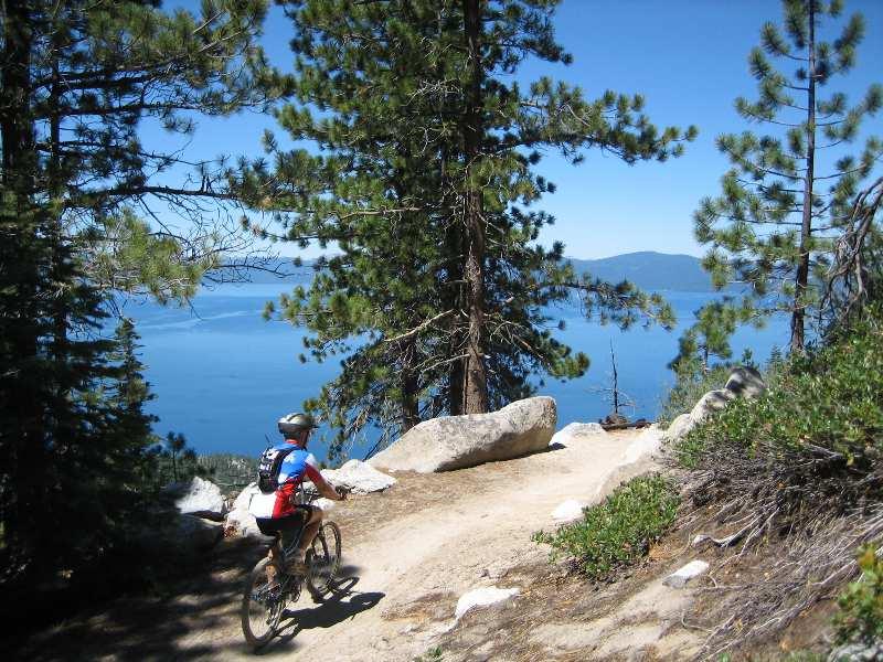 A mountain biker rides along a dirt trail surrounded by coniferous trees, with a stunning view of a lake and mountains in the background under a clear blue sky. Flume Trail mountain bike trail.