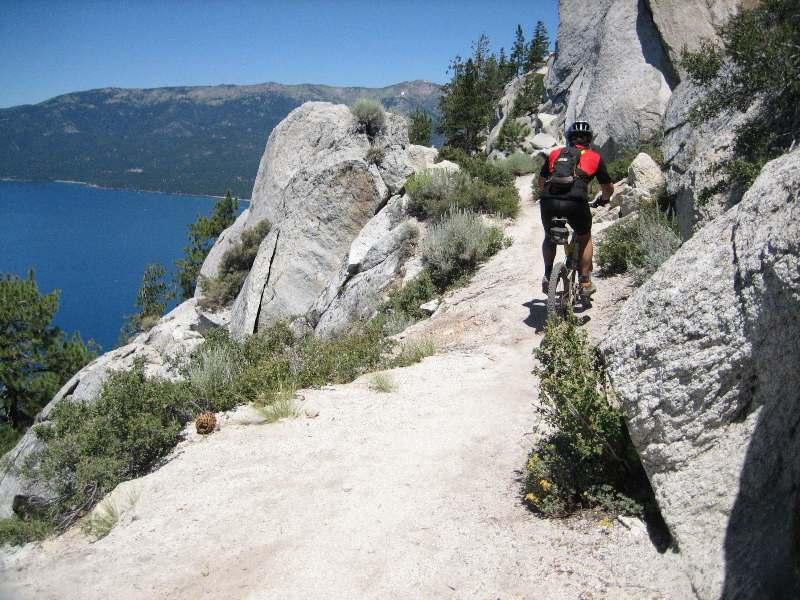 Biker riding along a narrow trail bordered by large rocks and shrubs, with a view of a blue lake and mountains in the background on a sunny day. Flume Trail mountain bike trail.