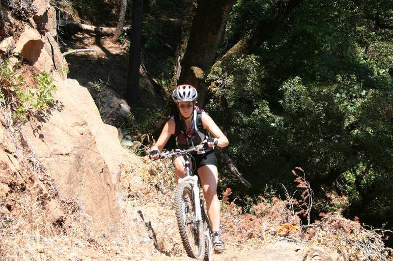 A person riding a mountain bike uphill on a rocky trail surrounded by trees and greenery. The rider is wearing a helmet and athletic clothing, demonstrating effort as they navigate the uneven terrain. Flume Trail mountain bike trail.