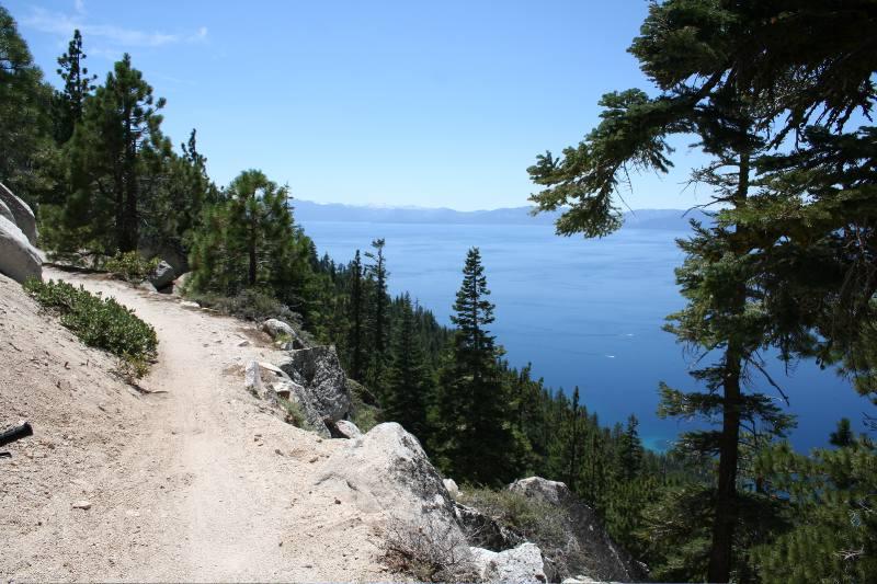 A sandy hiking trail winds through lush green pine trees, offering a scenic view of a blue lake and distant mountains under a clear blue sky. Flume Trail mountain bike trail.