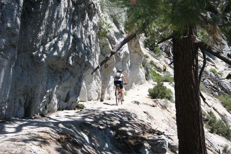 A cyclist riding on a narrow trail surrounded by rocky cliffs and greenery on a sunny day. Flume Trail mountain bike trail.