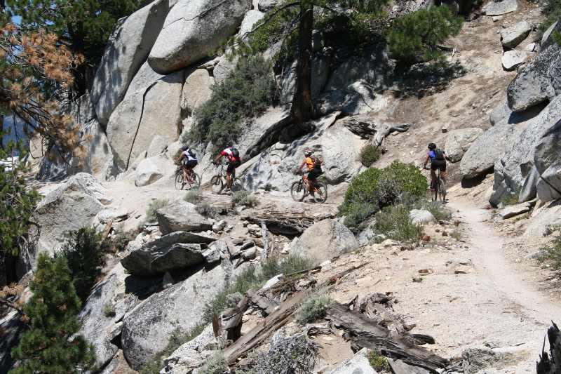 Four mountain bikers navigate a rocky trail surrounded by large boulders and sparse vegetation. The sunlit scene features a winding path that leads through natural terrain, showcasing the outdoor adventure in a rugged landscape. Flume Trail mountain bike trail.