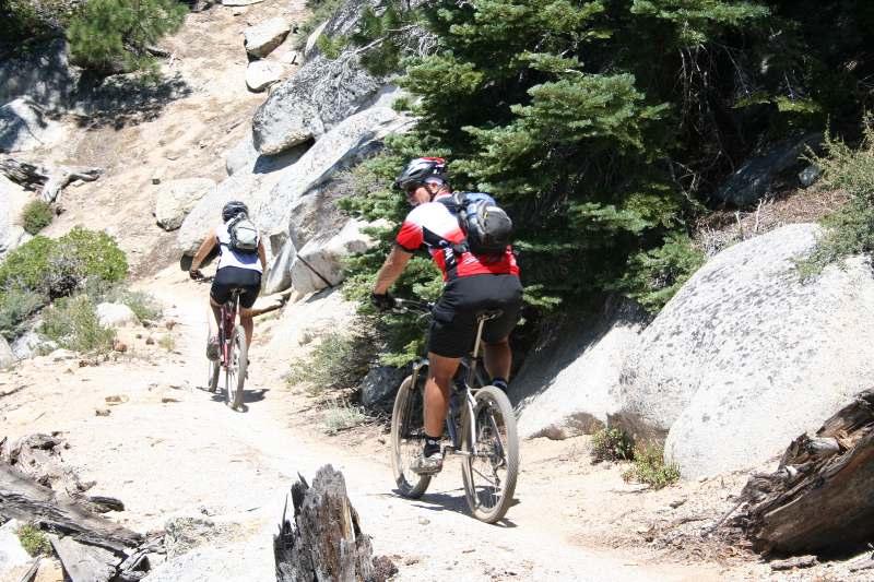 Two mountain bikers riding along a rocky trail surrounded by trees and boulders on a sunny day. Flume Trail mountain bike trail.