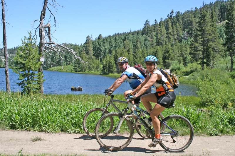 Two mountain bikers riding on a dirt path next to a lake, surrounded by green grass and trees. The first rider, wearing a blue helmet and a blue shirt, looks back while pedaling, and the second rider, wearing an orange and black outfit, smiles at the camera. The background features lush hills and a clear blue sky. Flume Trail mountain bike trail.