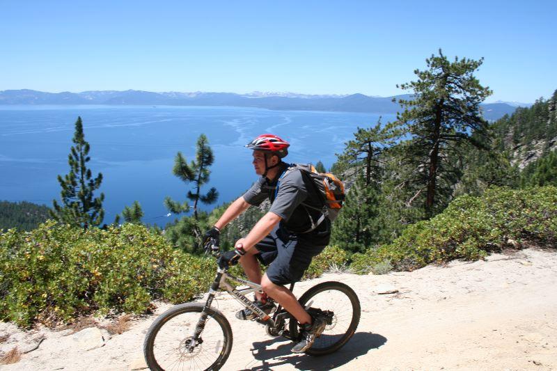 A mountain biker riding along a trail with a scenic view of a lake and mountains in the background. The sky is clear and blue, with pine trees lining the path. The biker is wearing a helmet and a backpack, showcasing an active outdoor lifestyle. Flume Trail mountain bike trail.