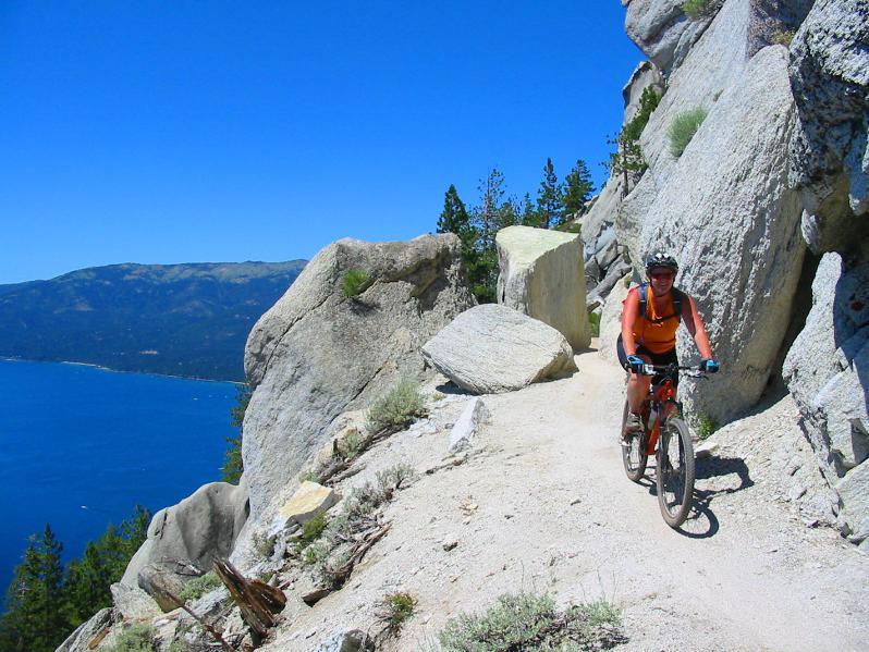 A mountain biker rides along a narrow dirt trail among large rocks, with a clear blue sky and a view of a lake and mountains in the background. Flume Trail mountain bike trail.