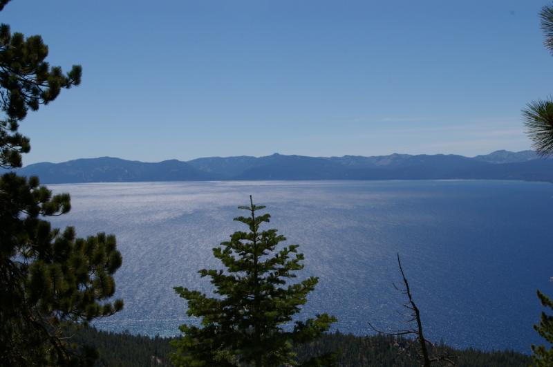 A panoramic view of a tranquil lake surrounded by mountains under a clear blue sky. The surface of the water reflects the sunlight, creating a shimmering effect. In the foreground, a few green trees frame the scene. Flume Trail mountain bike trail.