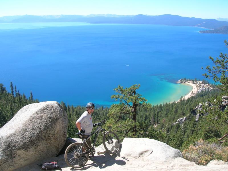 A person stands next to a mountain bike on a rocky outcrop, overlooking a scenic view of a clear blue lake surrounded by forested hills and distant mountains. The sun shines brightly in a clear sky. Flume Trail mountain bike trail.