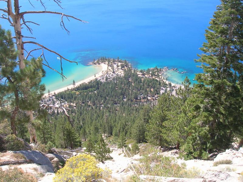 Aerial view of a serene coastal landscape featuring a sandy beach surrounded by dense green forests and clear blue water. The scene captures the contrast between the vibrant turquoise sea and the lush vegetation, offering a glimpse of a tranquil seaside community nestled along the shoreline. Flume Trail mountain bike trail.