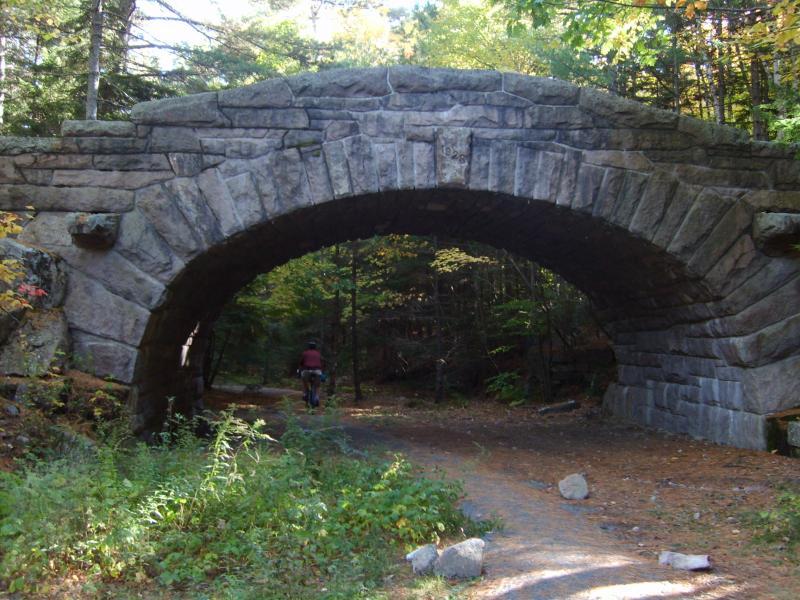 A stone arch bridge over a dirt path surrounded by dense trees and foliage. A person riding a bicycle can be seen approaching the bridge, with colorful autumn leaves visible in the background. Acadia Carriage Roads mountain bike trail.