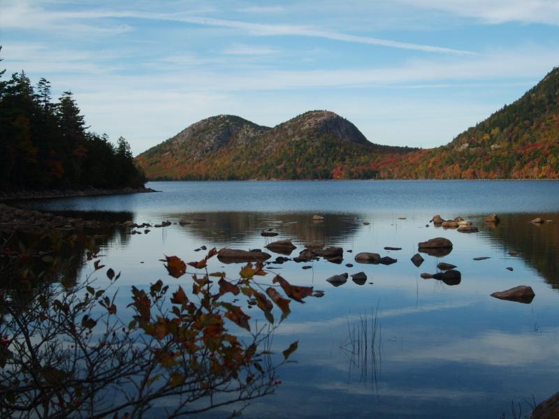 A tranquil lake scene featuring two rounded hills in the background, surrounded by colorful autumn foliage. In the foreground, rocks are visible in the water, with reflections on the lake's surface. The sky is clear with light clouds. Acadia Carriage Roads mountain bike trail.