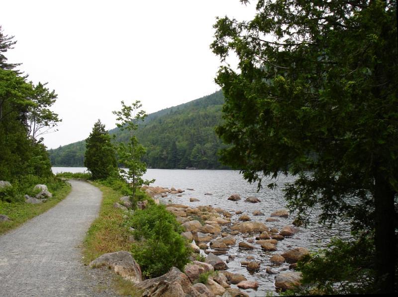 A scenic path lined with greenery meanders alongside a calm lake, bordered by rocky shores and rolling hills in the background. The sky is overcast, enhancing the tranquil atmosphere of this natural setting. Acadia Carriage Roads mountain bike trail.