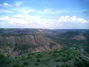 A panoramic view of a canyon landscape under a partly cloudy sky, featuring layered rock formations and sparse vegetation on the canyon floor. Palo Duro Canyon mountain bike trail.