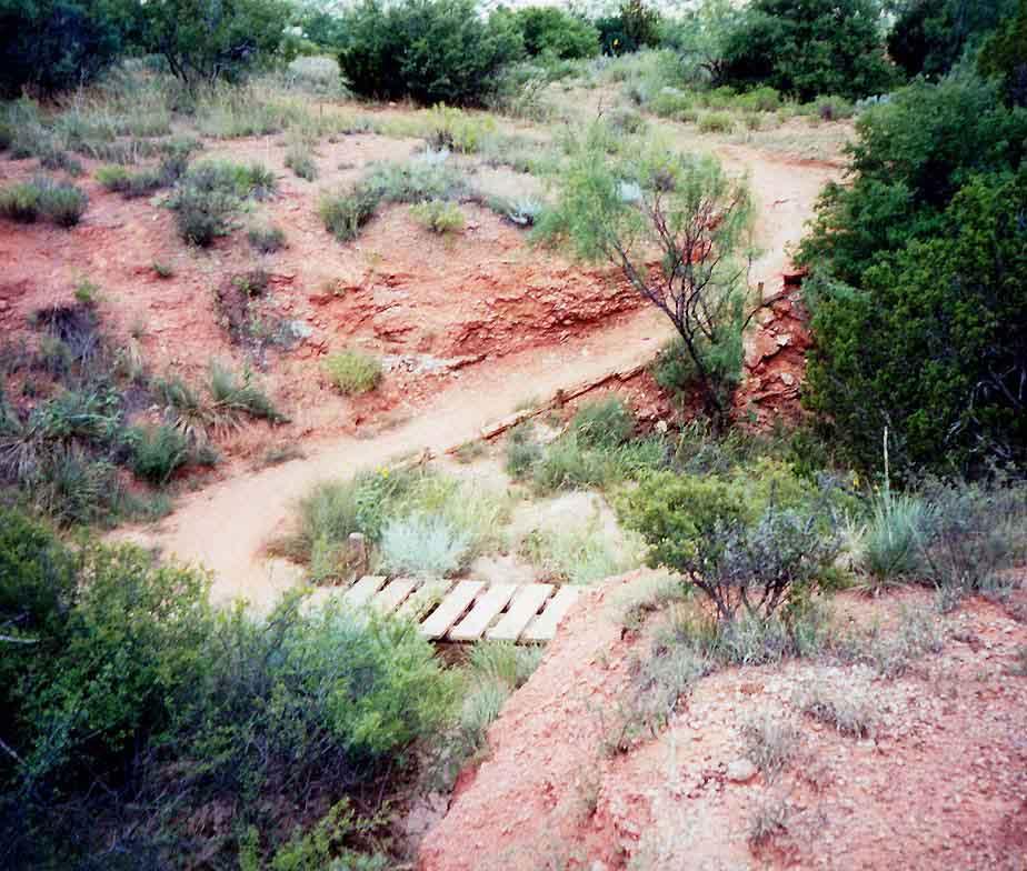 A winding dirt trail through a rocky landscape with reddish soil, flanked by sparse vegetation and small bushes. A wooden bridge crosses a small dip in the terrain, leading the way along the path. Palo Duro Canyon mountain bike trail.