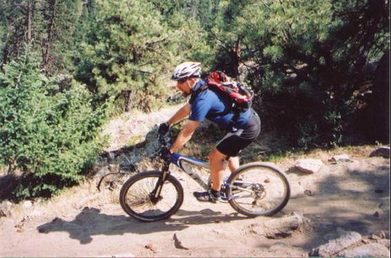 A mountain biker riding on a dirt trail surrounded by trees, wearing a helmet and a backpack, with a focus on the rocky terrain. Walker Ranch mountain bike trail.