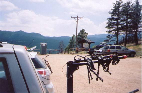 A parking area in a mountainous landscape, featuring several cars and a bike rack with bicycles. A wooden shelter and portable restrooms are visible in the background, with trees and mountains under a cloudy sky. Walker Ranch mountain bike trail.