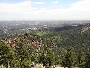 A panoramic view of a mountainous landscape featuring lush green trees and rocky outcrops, with a valley and distant plains visible under a blue sky with scattered clouds. Walker Ranch mountain bike trail.