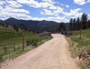 A dirt road winding through a grassy landscape, flanked by trees and mountains in the background under a partly cloudy blue sky. Walker Ranch mountain bike trail.