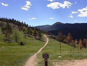 A winding dirt path through a green landscape, with scattered trees and mountains in the background under a blue sky with fluffy clouds. A wooden sign is visible near the path. Walker Ranch mountain bike trail.