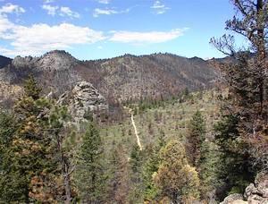 A scenic mountain landscape featuring rocky outcrops, dense coniferous forests, and a winding dirt path leading through the greenery under a partly cloudy sky. Walker Ranch mountain bike trail.