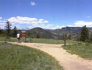 A cyclist standing near a trail sign in a grassy area, with mountains and a clear blue sky in the background. Walker Ranch mountain bike trail.
