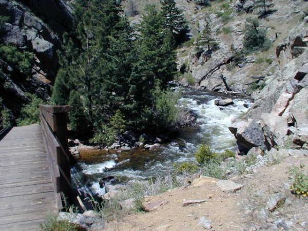 A wooden bridge overlooking a rushing river surrounded by rocky terrain and lush green trees. The scene captures the natural beauty of a mountainous area with flowing water and a variety of vegetation. Walker Ranch mountain bike trail.