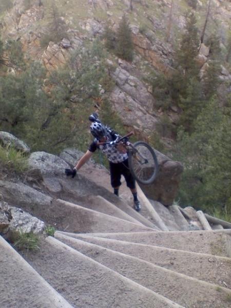 A cyclist climbing steep concrete stairs while carrying a mountain bike on one shoulder, surrounded by rocky terrain and trees in a natural setting. Walker Ranch mountain bike trail.