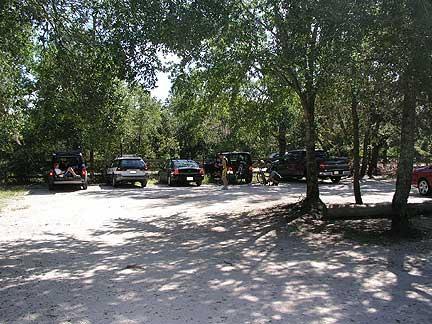 A parking area surrounded by trees, featuring several cars parked on a dirt lot. Sunlight filters through the foliage, casting shadows on the ground. Little Big Econ State Forest mountain bike trail.