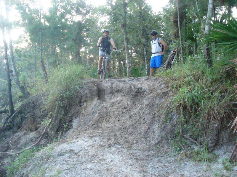 Two men are in a wooded area, one on a mountain bike at the edge of a dirt ramp, preparing to ride down, while the other stands nearby, observing. The surroundings are lush with trees and tall grass, and the sunlight filters through the foliage. Little Big Econ State Forest mountain bike trail.