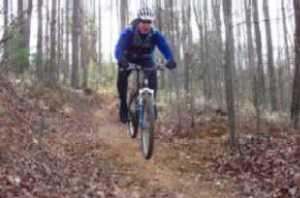 A person riding a mountain bike on a dirt trail surrounded by trees, wearing a helmet and blue jacket. The scene captures the essence of outdoor cycling in a forested area. Dark Mountain Trail mountain bike trail.