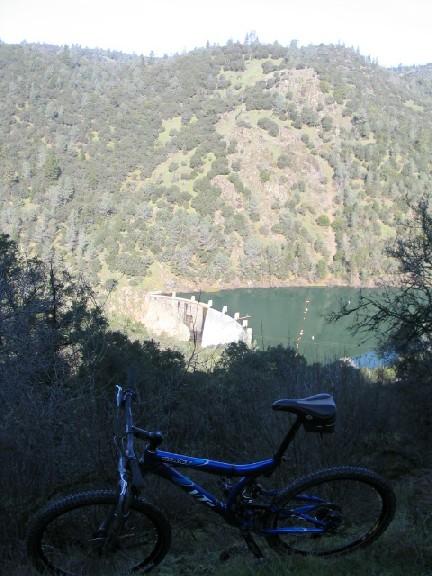 A mountain bike is positioned in the foreground, with a scenic view of a lake and green hills in the background. A dam is visible across the water, surrounded by lush vegetation under a clear blue sky. Clementine Loop mountain bike trail.