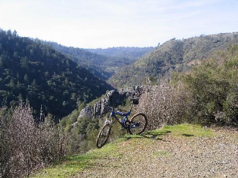 A blue mountain bike rests on a grassy trail overlooking a lush valley surrounded by hills and trees, under a clear sky. Clementine Loop mountain bike trail.